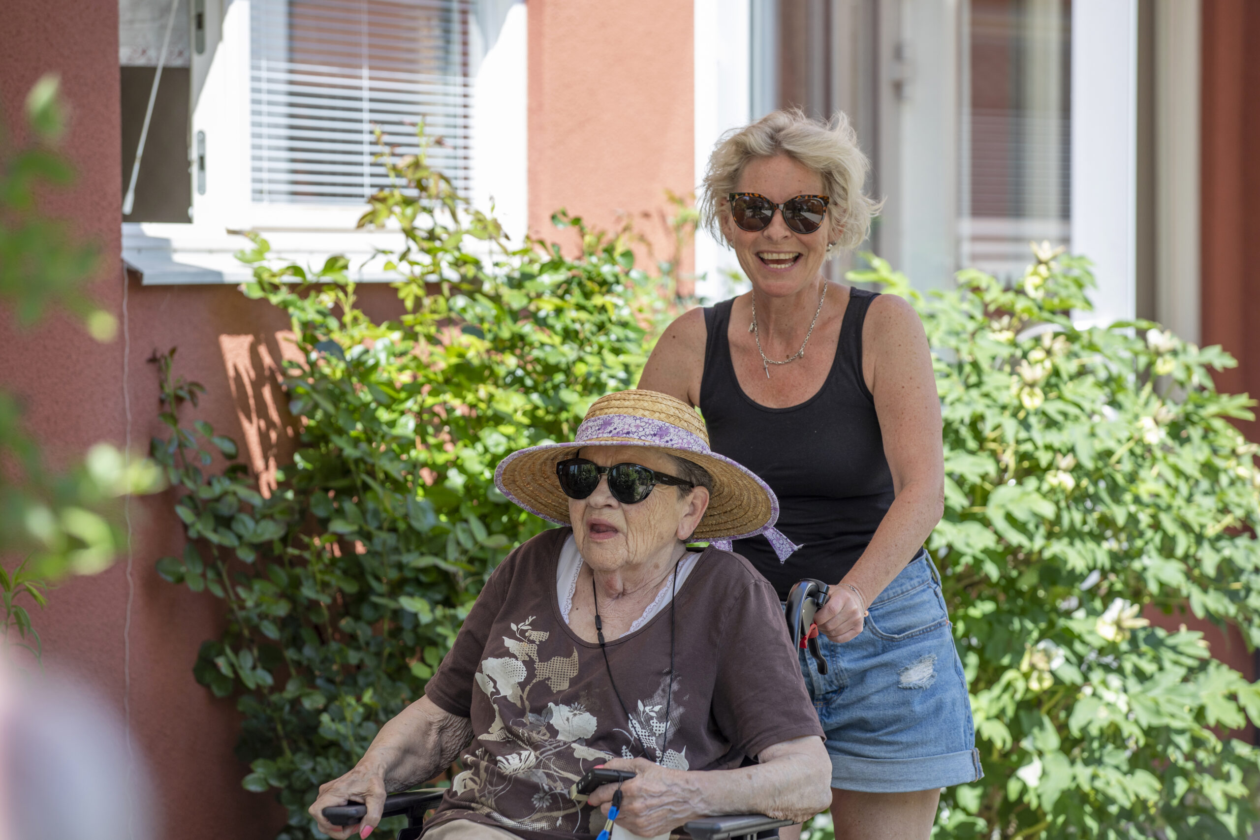 Kvinna skjutser äldre kvinna i rullstol en lummig sommardag.