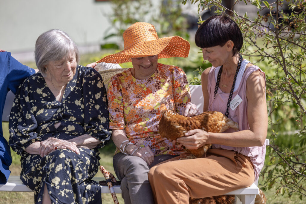 Två äldre kvinnor i blommiga sommarkläder vid yngre kvinna med höna i knäet.